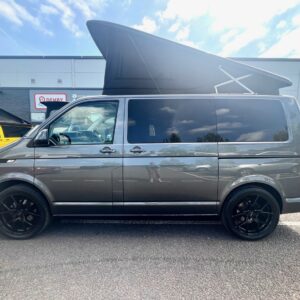 An Indium Grey SWB T6.1 Volkswagen Transporter Highline Campervan (73 plate, A1433) with a raised pop-top roof is parked in a lot on a sunny day, with a yellow van visible near a modern building in the background.