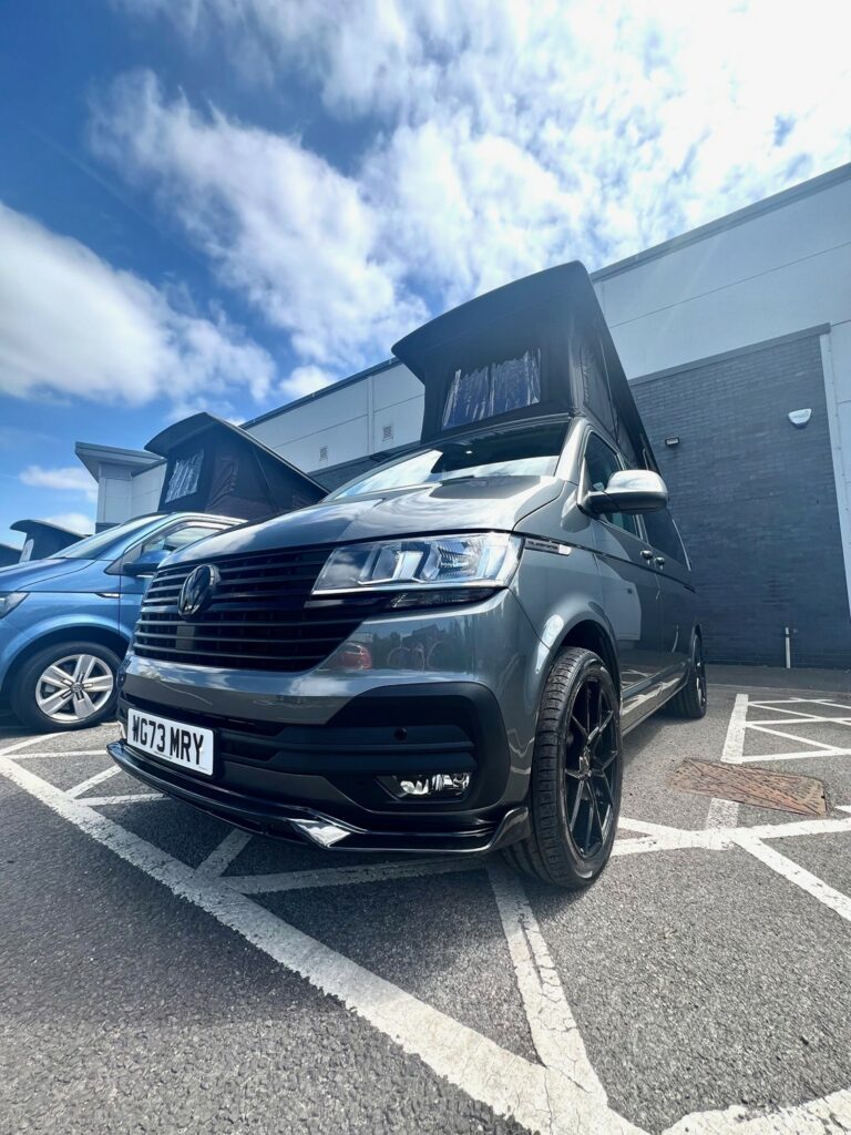 An Indium Grey SWB T6.1 Volkswagen Transporter Highline Campervan (73 plate, A1433) is parked in a marked space outside a building on a sunny day, showing its front view and license plate WG73 WRY.