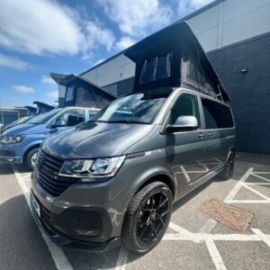An Indium Grey SWB T6.1 Volkswagen Transporter Highline Campervan (73 plate, A1433) with a pop-up roof is parked in a lot under partly cloudy skies, with another similar van visible in the background.