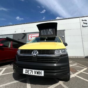 A SWB T6.1 Volkswagen Transporter Startline Campervan in Sunny Yellow (71 Plate, A1441) is parked outside a large white and gray building under a blue sky with clouds.