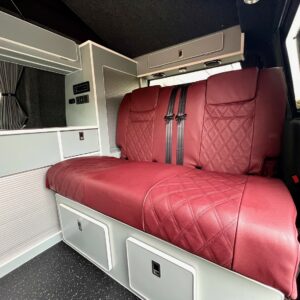 The SWB T6.1 Volkswagen Transporter Highline Campervan (Fortana Red, 23 Plate, A1443) features a red leather bench seat with seatbelts and diamond stitching, white storage cabinets, and a black speckled floor.