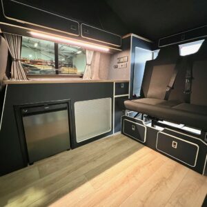 The SWB T6.1 Volkswagen Transporter Highline Campervan (Indium Grey, 73 plate, A1438) features light wood flooring, black seating with storage, a mini fridge, window with curtains, and overhead cabinets. Workshop view through window.