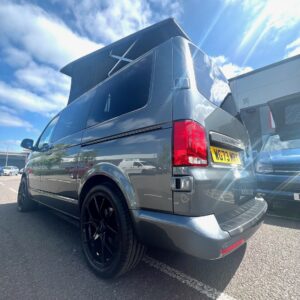 An Indium Grey SWB T6.1 Volkswagen Transporter Highline Campervan (73 plate, A1433) with a raised pop-top roof and black wheels is parked in a lot on a sunny day, with other cars and a building in the background.