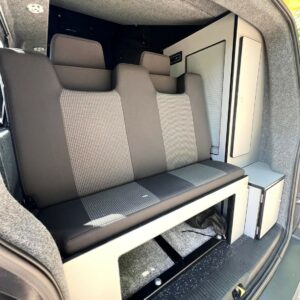 The SWB T6.1 Volkswagen Transporter Startline Campervan (Sunny Yellow, 71 Plate, A1441) features a gray and black upholstered bench seat, storage cabinets, and gray carpeted walls seen from the side door—a clean, modern space for travel or camping.