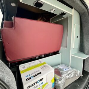The image shows the interior of a Fortana Red SWB T6.1 Volkswagen Transporter Highline Campervan (23 Plate, A1443) with a folded maroon seat, built-in light blue storage units, Thetford Porta Potti box, and silver insulated floor mat.