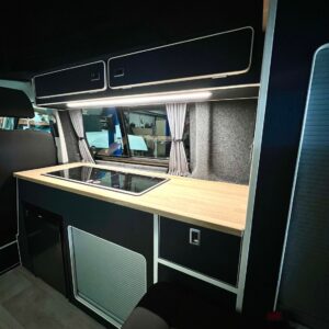 The SWB T6.1 Volkswagen Transporter Highline Campervan (Indium Grey, 73 plate, A1438) features a modern kitchen with wooden countertop, built-in stove, black and white trimmed cabinets, gray-curtained window, and bright LED lighting.