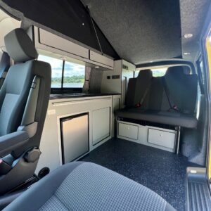 The SWB T6.1 Volkswagen Transporter Startline Campervan in Sunny Yellow (71 Plate, A1441) features folded bench seats, a compact kitchenette with cabinets and fridge, swivel front seats, and windows for natural light in a clean, organized interior.