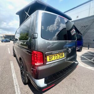 An Indium Grey SWB T6.1 Volkswagen Transporter Highline Campervan (73 plate, A1438) with the roof extended is parked in a lot on a sunny day. Other cars and a building are visible in the background. The license plate reads DF73 ZHR.