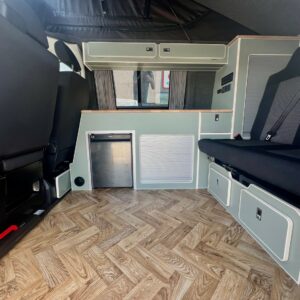 The SWB T6.1 Volkswagen Transporter Highline Campervan (Indium Grey, 73 plate, A1433) features light green cabinets, wood-patterned vinyl flooring, black cushioned seats, ample storage, and bright natural light through the windows.