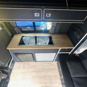 The SWB T6.1 Volkswagen Transporter Highline Campervan (Indium Grey, 73 plate, A1438) features a compact kitchen with wooden countertop, covered sink, upper cabinets, small fridge, seating area, window view of the front exterior, and light wood flooring.