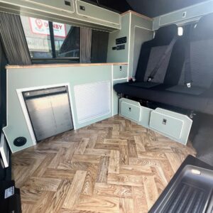 Interior of the SWB - T6.1 Volkswagen Transporter Highline Campervan (Indium Grey, 73 plate – A1433), featuring wood-patterned flooring, built-in storage, a fold-out bench seat, pale green cabinetry, and curtained windows.