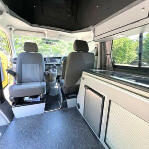 The SWB T6.1 Volkswagen Transporter Startline Campervan (Sunny Yellow, 71 Plate, A1441) features two front seats, a compact kitchenette with stove and fridge, large windows for sunlight and greenery views, and a partially covered ceiling.