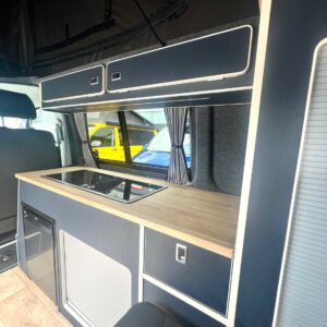 The SWB T6.1 Volkswagen Transporter Highline Campervan (Indium Grey, 73 plate, A1438) features a modern interior with a compact kitchen—wooden countertop, built-in stove, fridge, cabinets, and a window with curtains in sleek contrasting colors.