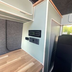 The SWB T6.1 Volkswagen Transporter Highline Campervan (Indium Grey, 73 plate, A1433) features a compact interior with wood countertop, light green cabinets, control panel, power outlets, a ribbed sliding door, and partial black seat on the right.
