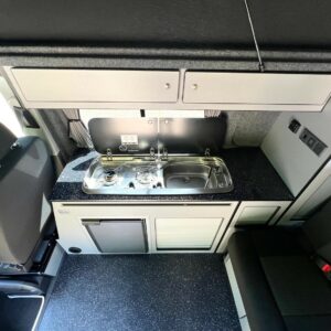 Overhead view of the SWB T6.1 Volkswagen Transporter Startline Campervan in Sunny Yellow (71 Plate, A1441), showing a compact kitchen with a small fridge, two-burner stove, sink with closed cover, white cabinets, and black countertops by the driver’s seat.