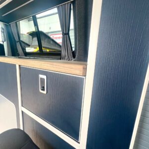 Interior view of a SWB - T6.1 Volkswagen Transporter Highline Campervan in Indium Grey (73 plate, A1438) showing dark blue wood paneling, a curtained window with silver handle, part of a seat, and a Denby sign outside.