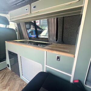 The SWB T6.1 Volkswagen Transporter Highline Campervan (Indium Grey, 73 plate, A1433) has a modern interior with light green cabinetry, a wooden countertop and stovetop, overhead cabinets, curtained window, wood-patterned floor, and black seat.