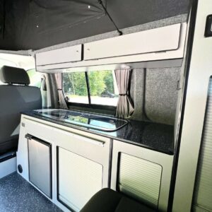 The SWB T6.1 Volkswagen Transporter Startline Campervan in Sunny Yellow (71 Plate, A1441) features a modern interior with a compact kitchenette—stove, sink, cabinets, drawers, fridge—and sunlight streaming through curtained windows.