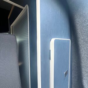 A close-up of the SWB - T6.1 Volkswagen Transporter Highline Campervan (Indium Grey, 73 plate, A1438) shows a modern blue cabinet with a vertical door, silver handle, and ribbed sliding panel against a gray carpeted wall.