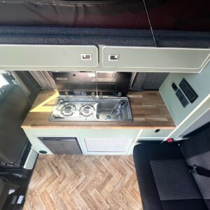 The SWB - T6.1 Volkswagen Transporter Highline Campervan (Indium Grey, 73 plate, A1433) features a compact kitchenette with a wooden countertop, built-in two-burner gas stove, stainless steel sink, overhead cabinets, and adjacent seating area.