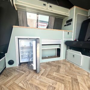 The SWB T6.1 Volkswagen Transporter Highline Campervan (Indium Grey, 73 plate A1433) features light wood herringbone flooring, an open small fridge, built-in cabinets, a folded bench seat, and a window showing a Denby sign outside.
