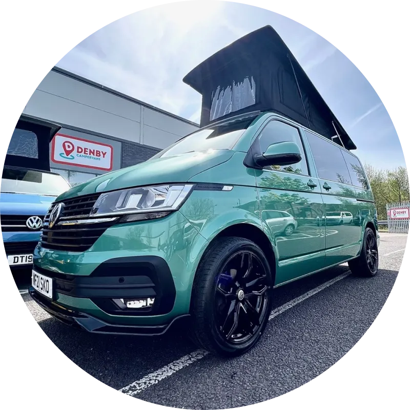 A green Volkswagen camper van with a pop-up roof is parked in a lot outside a Denby dealership, with black alloy wheels and another blue van beside it, under a partly cloudy sky.