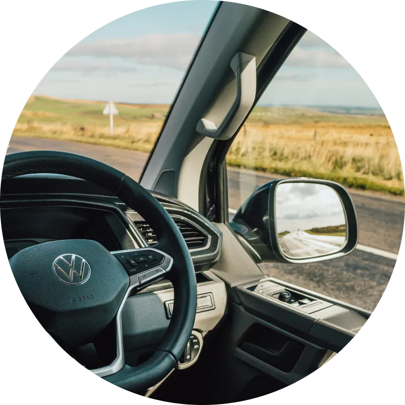 View from inside a Volkswagen vehicle, showing the steering wheel, side mirror, and part of the dashboard, with an empty road and grassy fields visible through the window.