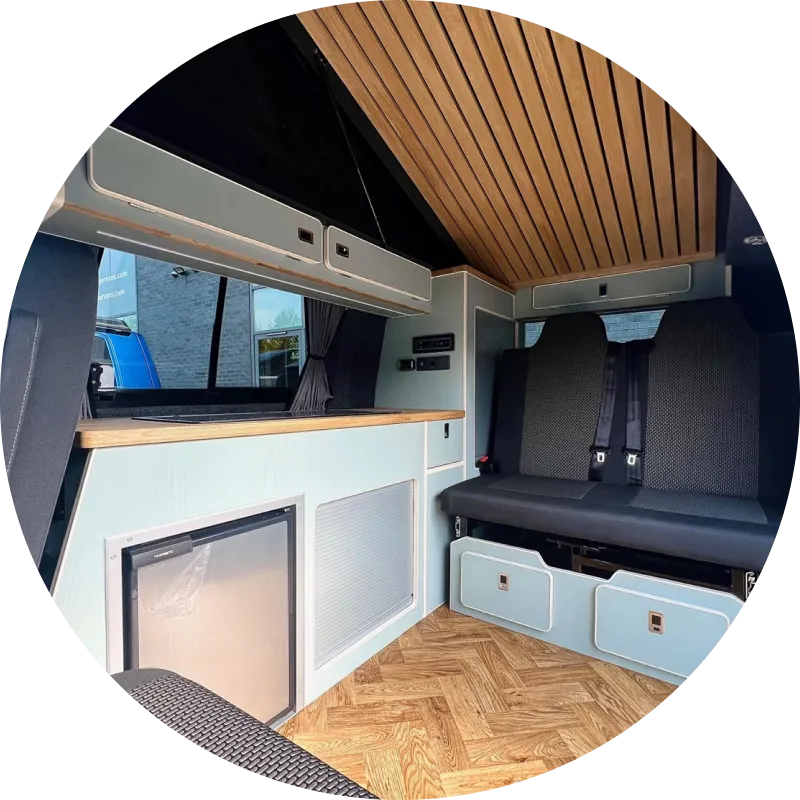 Modern camper van interior with wood panel ceiling, built-in seating with storage drawers, a small counter with window above, and compact fridge below, all in a clean and minimalist design.