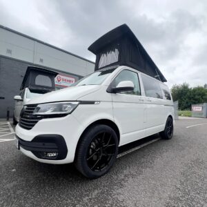 A Candy White SWB T6.1 Volkswagen Transporter Highline Campervan (24 plate, A1447) with a raised black pop-top roof and black wheels is parked in a lot near a building under an overcast sky; DENBY sign visible in background.