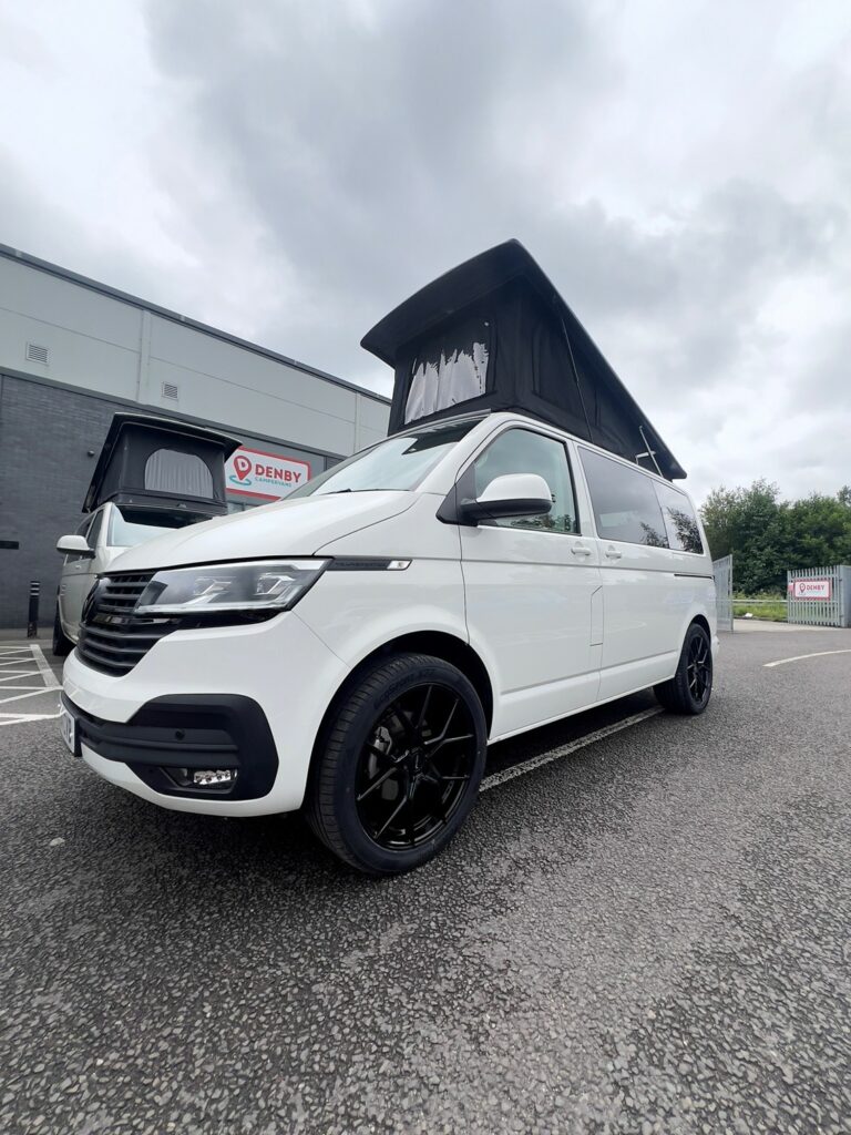 A Candy White SWB T6.1 Volkswagen Transporter Highline Campervan (24 plate, A1447) with a raised black pop-top roof and black wheels is parked in a lot near a building under an overcast sky; DENBY sign visible in background.