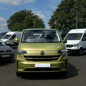 A Warm Green 2025 ALL-NEW Volkswagen Transporter T7 DSG Auto Commerce Plus Campervan (Deluxe SWB, Stock A1449) is parked in a lot among white vans and trees on a sunny day, facing the camera.