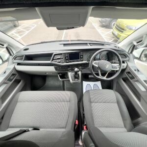Interior view of the SWB T6.1 Volkswagen Transporter Highline Campervan (Candy White, 24 plate, A1447) from the back seat, showing two gray front seats, right-side steering wheel, dashboard with touchscreen, gear stick, and floor mats.