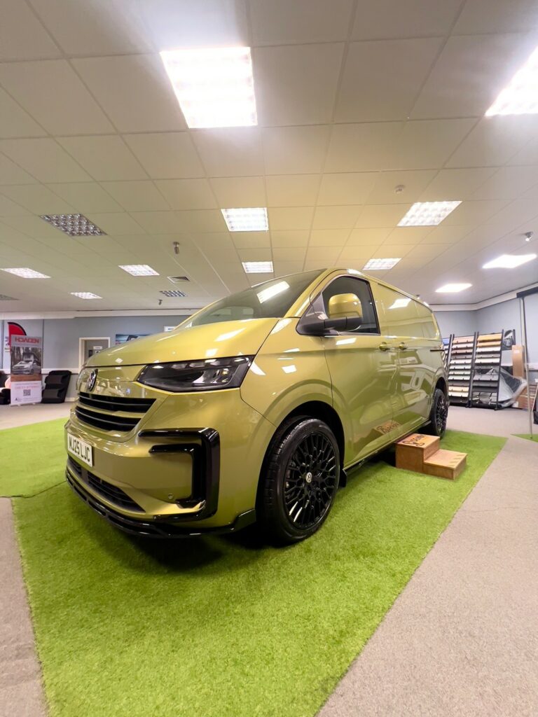 A Deluxe SWB - 2025 ALL-NEW Volkswagen Transporter T7 DSG Auto Commerce Plus Campervan in Warm Green (Stock A1449) is displayed indoors on artificial grass with wooden steps, office furniture, and shelves visible under bright ceiling lights.