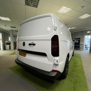 A 2025 ALL-NEW Volkswagen Transporter T7 150BHP Manual Commerce Plus Campervan in Clear White (Stock A1444) is parked indoors on green artificial grass under fluorescent office lights, featuring tinted windows and a yellow license plate.
