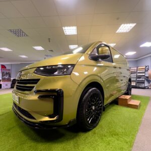 The 2025 ALL-NEW Volkswagen Transporter T7 DSG Auto Commerce Plus Campervan in Warm Green (Deluxe SWB, Stock A1449) is showcased indoors on artificial grass under bright lights, with black wheels, a wooden step, and display banners nearby.
