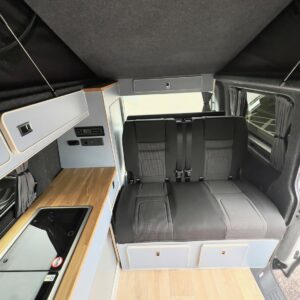The SWB T6.1 Volkswagen Transporter Highline Campervan (Candy White, 24 plate, A1447) features a compact wooden kitchenette with sink and stove on the left, plus a black cushioned bench with seatbelts on a light wood floor.