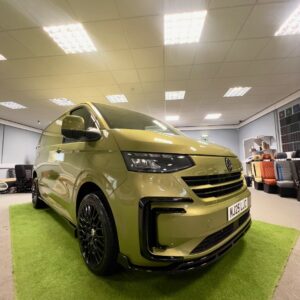 A 2025 ALL-NEW Volkswagen Transporter T7 DSG Auto Commerce Plus Campervan in Warm Green (Deluxe SWB, Stock A1449) is parked indoors on artificial grass, surrounded by office furniture under bright ceiling lights.