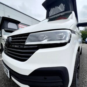 Close-up of the front of a Candy White SWB T6.1 Volkswagen Transporter Highline Campervan (24 plate, A1447) with its roof extended, parked on tarmac with another similar van and a building in the background.
