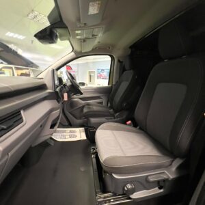 The image shows the interior of a Deluxe SWB - 2025 ALL-NEW Volkswagen Transporter T7 DSG Auto Commerce Plus Campervan in Warm Green, with two dark upholstered front seats, dashboard controls, and a large windshield reflecting showroom lights.