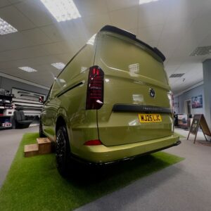A Deluxe SWB 2025 Volkswagen Transporter T7 DSG Auto Commerce Plus Campervan in Warm Green (Stock A1449) is displayed indoors on artificial grass, surrounded by automotive accessories and bright ceiling lights.