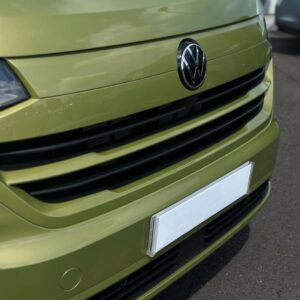 Close-up of the front of a Deluxe SWB - 2025 ALL-NEW Volkswagen Transporter T7 DSG Auto Commerce Plus Campervan in Warm Green, showing the VW logo on the grill and a blank white license plate, parked on asphalt. Stock A1449.