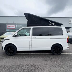 A Candy White SWB T6.1 Volkswagen Transporter Highline Campervan (24 plate, A1447) with a black pop-up roof, black wheels, and tinted windows is parked outside a gray industrial building on a cloudy day.