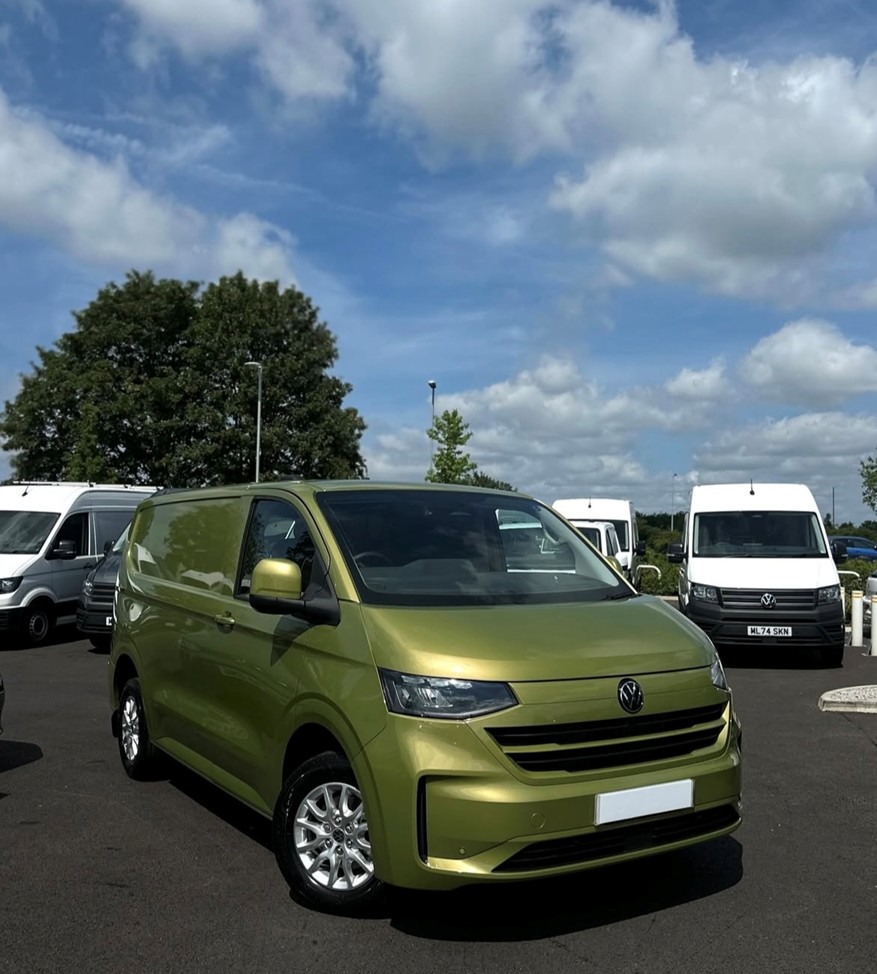 A Deluxe SWB 2025 ALL-NEW Volkswagen Transporter T7 DSG Auto Commerce Plus Campervan in Warm Green (Stock A1449) is parked in a lot with several white vans behind it, all beneath a partly cloudy blue sky.