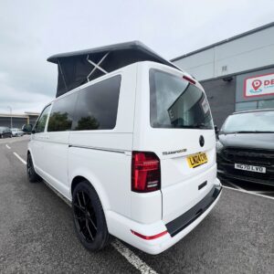 A Candy White SWB T6.1 Volkswagen Transporter Highline Campervan (24 plate, A1447) with black wheels and a raised pop-top roof is parked near a gray building next to a black car.