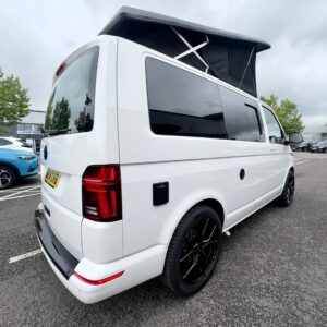 A Candy White SWB T6.1 Volkswagen Transporter Highline campervan (24 plate, A1447) with a raised pop-up roof, black-tinted windows, and black rims is parked in a lot under an overcast sky, with other vehicles and trees in the background.
