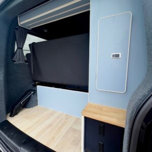 The SWB T6.1 Volkswagen Transporter Highline Campervan (Candy White, 24 plate, A1447) features light wood flooring, blue/white cabinetry, a foldable black seat/bed, compact counter, storage drawers, gray walls, and curtained window.