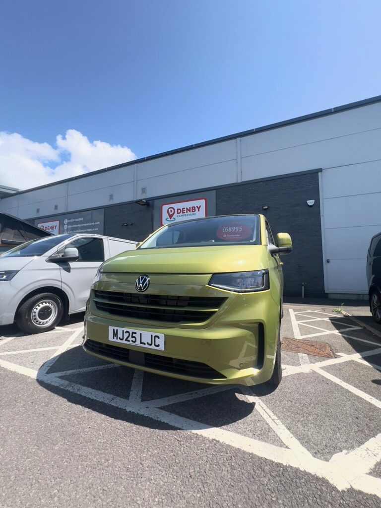 A Deluxe SWB - 2025 ALL-NEW Volkswagen Transporter T7 DSG Auto Commerce Plus Campervan in Warm Green (Stock A1449) is parked by a building with Denby signs, displaying a £48,995 price tag. Other vehicles are nearby under a sunny sky.