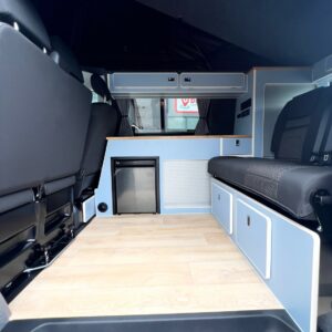 Interior of the SWB T6.1 Volkswagen Transporter Highline Campervan (Candy White, 24 plate, A1447) with light wood floors, compact fridge, light blue cabinets, foldable bench seats, and black front seats. Modern and organized space.