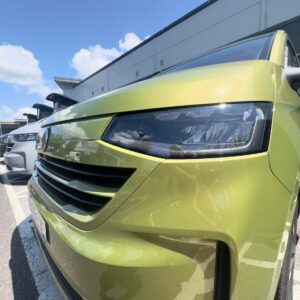 Close-up of the 2025 ALL-NEW Volkswagen Transporter T7 DSG Auto Commerce Plus Campervan in Warm Green (Stock A1449), parked outdoors with blue sky reflecting on its hood; other vehicles and a building appear in the background.