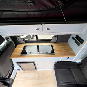 Interior view of the SWB - T6.1 Volkswagen Transporter Highline Campervan (Candy White, 24 plate, A1447) featuring a wooden countertop, two covered sink/stove areas, upper/lower storage cabinets, light wood flooring, and a black seat on the right.